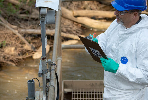 Man wearing a hard hat and Tyvek suit taking notes on a clipboard as he studies equipment outside.